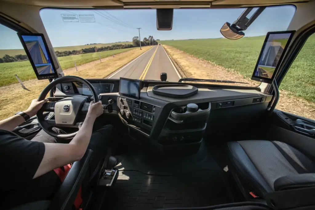 Interior da cabine em viagem: as mãos do motorista no volante e a rodovia reta à frente, entre campos abertos.